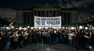 Fotografía cinematográfica nocturna de una multitud frente a una universidad pública argentina. Cientos de estudiantes y docentes sostienen libros abiertos que emiten una intensa luz blanca, iluminando una plaza oscura. En el centro destaca una pancarta con el texto "LAS UNIVERSIDADES NO SE APAGAN"