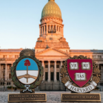 Fotografía de la fachada del Congreso de la Nación Argentina durante el atardecer, con el Escudo Nacional Argentino y el logo de la Universidad de Harvard en primer plano sobre pedestales metálicos.